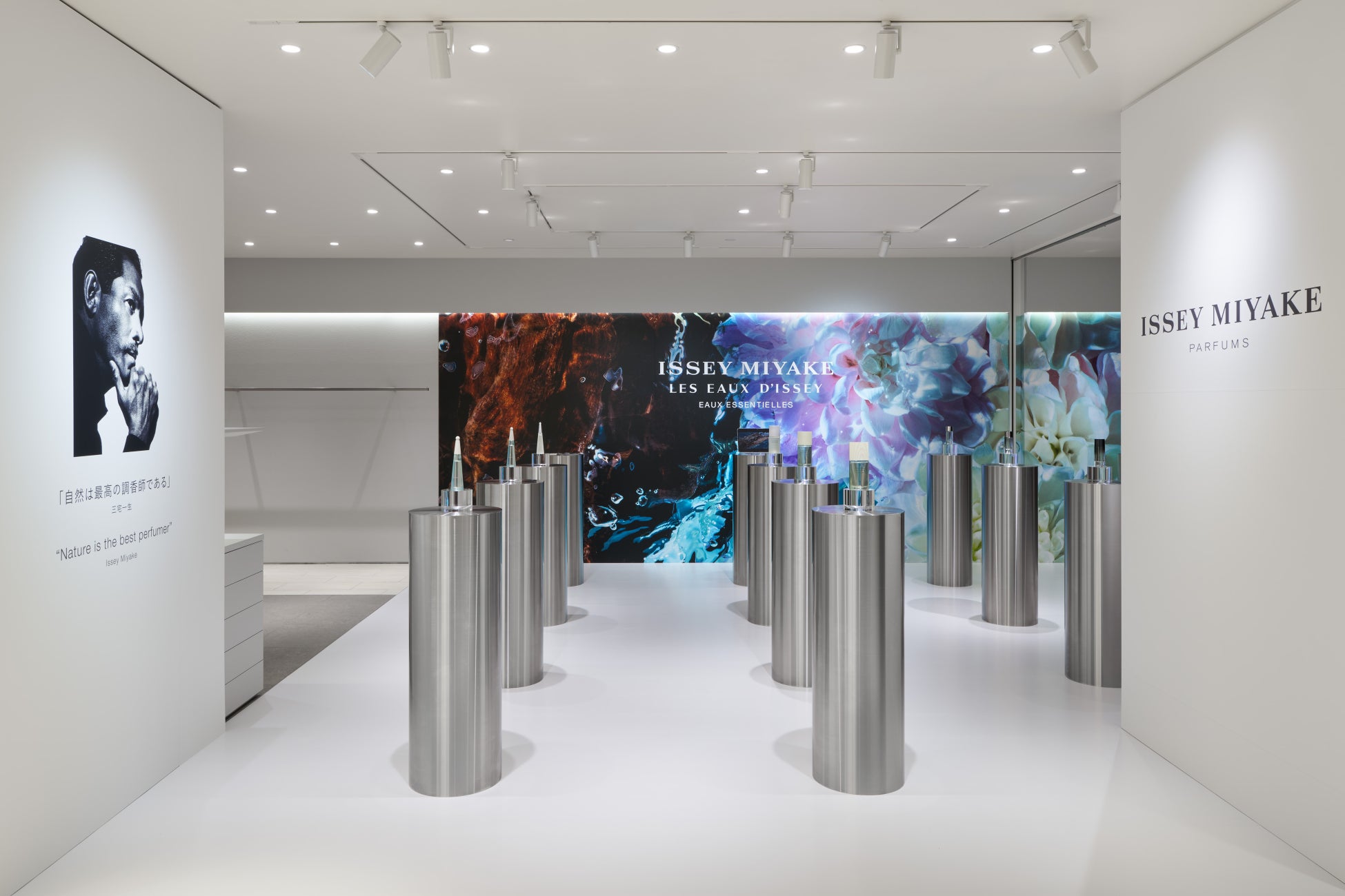 銀色の円柱什器に香水ボトルが並ぶ白い展示空間。奥の壁面に花や水を思わせる図柄、左側に人物写真と説明文が掲示されている。